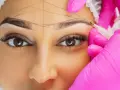 Nahaufnahme einer Frau, deren Augenbrauen mit Linien markiert werden, während eine Person in pinken Handschuhen mit einem Stift arbeitet.
